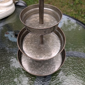 May include: A three-tiered metal serving stand with a galvanized finish. The stand features a central vertical rod topped with a spherical finial, connecting three circular trays of decreasing size from bottom to top, all with a textured surface.