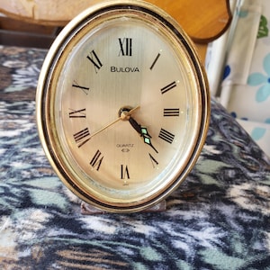 Op de afbeelding: Een ovale Bulova kwartsklok met een goudkleurige wijzerplaat, zwarte Romeinse cijfers als uurmarkeringen en dunne zwarte wijzers. De klok heeft een metallic gouden afwerking op de rand en het woord Bulova in het zwart op de wijzerplaat.