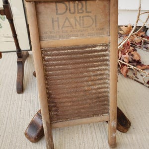 Op de afbeelding: Antieke houten wasbord met de tekst "DUBL HANDI" en "COLUMBUS WASHBOARD CO. COLUMBUS, O". Het wasbord heeft een gegolfd metalen schuuroppervlak en een houten frame. Een vintage wasgoedgereedschap.