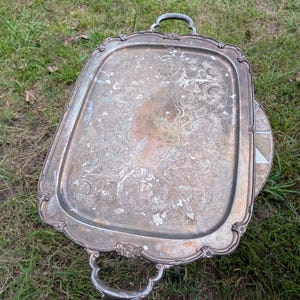 May include: A rectangular silver-toned serving tray with two ornate handles and a decorative scalloped edge. The surface features an intricate, tarnished floral engraving in the centre, showing signs of wear and oxidation across the metal.