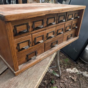 Può includere: Un mobiletto rettangolare in legno con quindici piccoli cassetti disposti su tre file. Ogni cassetto è realizzato in legno marrone chiaro ed è dotato di una maniglia in metallo scuro e di una cornice portatichetta sulla parte frontale.