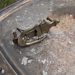 May include: A brass drawer pull with a curved, elongated shape and a central hinged handle. The hardware features a tarnished, metallic gold finish and includes two rusted metal screws attached to the back for mounting purposes.