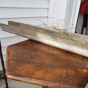 May include: An antique wooden table with a dark, distressed finish. A long, weathered wooden trough rests on the table. A white, metal decorative piece sits behind the trough. The table has a unique, octagonal shape.