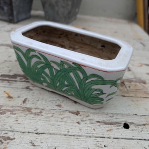 May include: A rectangular white ceramic planter featuring a hand-painted green grass motif along the exterior sides. The vessel has a smooth, glossy finish with a thin orange line accenting the upper rim and a hollow, unglazed interior basin.