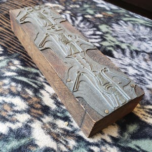 May include: A rectangular wooden block featuring a metallic printing plate attached to the top surface. The silver-toned metal plate displays intricate, raised relief patterns of stylized figures and geometric lines, secured by small metal pins.