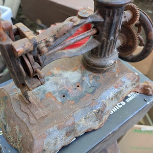 May include: A vintage, heavily rusted cast iron sewing machine with a weathered, dark metal finish. The machine features a prominent hand crank wheel with visible gears, a needle bar assembly, and remnants of decorative gold-toned filigree paint.