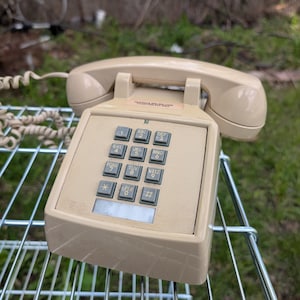 May include: A beige landline telephone featuring a rectangular base with a push-button keypad, including numbers zero through nine, an asterisk, and a pound sign. A matching beige handset rests on the cradle, connected by a coiled cord.