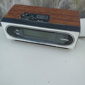 May include: A rectangular digital alarm clock with a wood grain top panel and a white front face. The display area features the text AM, PM, and COPAL. A black control panel on top includes an alarm switch and a snooze button with visible labels.