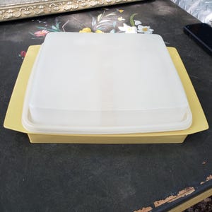 May include: A square, pale yellow plastic storage container with a matching translucent white lid. The container features wide, flat handles on two opposite sides and a smooth, flat surface on the top of the lid, providing a simple, functional design.