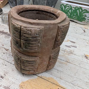 Puede incluir: Un recipiente cilíndrico de arcilla marrón con un borde ancho y grueso y un centro hueco. La superficie exterior está decorada con paneles rectangulares repetidos, cada uno con un patrón geométrico empotrado en un tono marrón oscuro.