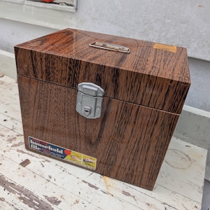 May include: A rectangular portable household file box with a brown faux wood grain finish. The box features a silver metal latch with a keyhole on the front, a silver metal handle on the top, and a label that reads Portable household file.