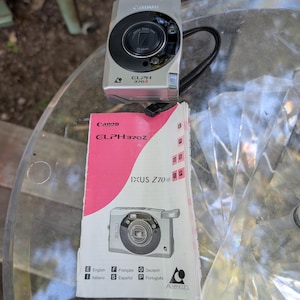 May include: A silver Canon ELPH 370Z compact camera with a black lens housing and a black wrist strap, accompanied by a white and pink instruction manual featuring the camera model name and the Advanced Photo System logo on the front cover.