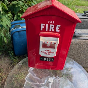 May include: A bright red plastic fire alarm pull station box with a peaked roof and a small red light on top. The front features white text reading FOR FIRE, OPEN, THEN PULL DOWN HOOK, and a black rectangular label with the white text 911.