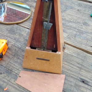 May include: A wooden metronome with a pyramid-shaped body, a central metal pendulum, and a vertical scale. The device features a light brown exterior, a darker wood panel behind the pendulum, and a detachable wooden front cover resting below.