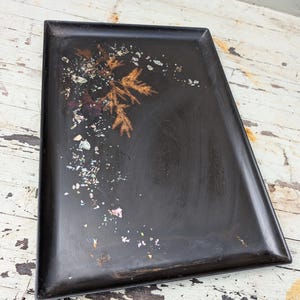 Peut inclure: Un plateau rectangulaire noir orné d'un motif incrusté de brindilles de pin brun doré et de paillettes irisées multicolores. La surface présente une finition sombre et lisse avec un rebord surélevé entourant le centre plat.