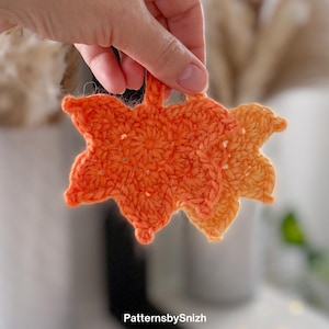 May include: Two crocheted orange and yellow autumn leaves. The leaves are held up in front of a blurred background. The leaves are made of yarn and have a textured appearance. The text "PatternsbySnizh" is visible in the bottom right corner.
