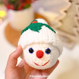 May include: A handmade, white crocheted snowman ornament with a green holly leaf and red berry accent on its hat. The snowman has blue eyes, a red nose, and a smiling red mouth. The text "PatternsbySnizh" is visible.