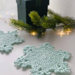 May include: Two light blue crocheted snowflake coasters. The coasters are on a white surface, with a dark green gift box and evergreen sprigs in the background. The coasters have a detailed, openwork design.