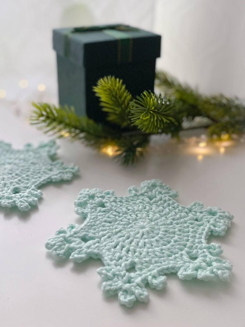 May include: Two light blue crocheted snowflake coasters. The coasters are on a white surface, with a dark green gift box and evergreen sprigs in the background. The coasters have a detailed, openwork design.