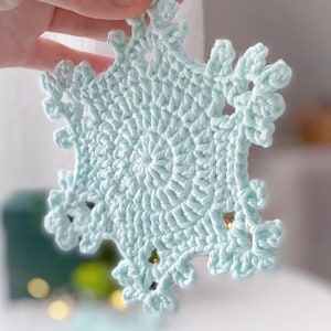 May include: A light blue, crocheted snowflake ornament. The snowflake is held up by a hand. The snowflake has a detailed, openwork design with a central circular pattern and radiating points. The ornament is likely handmade and suitable for winter holiday decor.