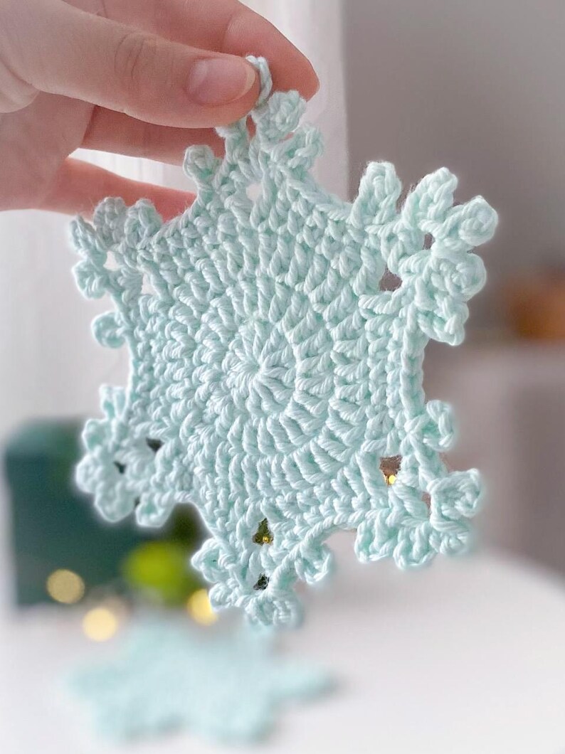 May include: A light blue, crocheted snowflake ornament. The snowflake is held up by a hand. The snowflake has a detailed, openwork design with a central circular pattern and radiating points. The ornament is likely handmade and suitable for winter holiday decor.