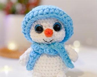 May include: A handmade crochet snowman with a blue hat and scarf. The snowman has large, black eyes, an orange nose, and a smiling mouth. The snowman is white with blue accents and is a winter-themed decoration.
