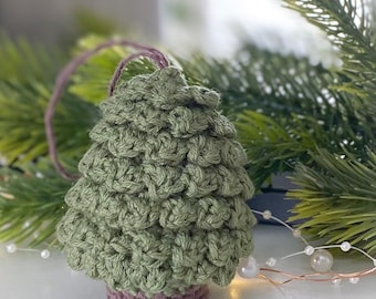 May include: A handmade, crocheted Christmas tree ornament. The tree is a textured green with a mauve base and hanging loop. The background includes evergreen branches and string lights.