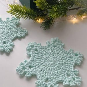 May include: Two light blue crocheted snowflake coasters. The coasters are handmade with a detailed openwork design. A green sprig of pine and a small green gift box are in the background, suggesting a holiday theme.