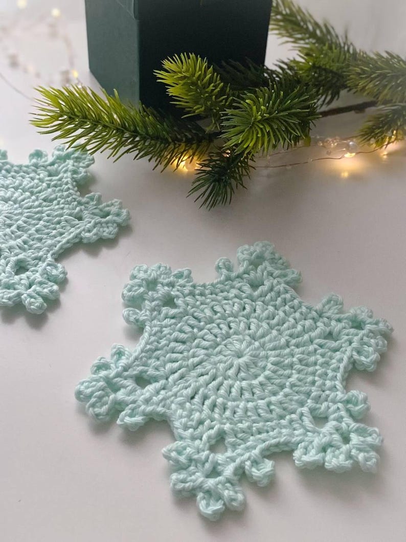 May include: Two light blue crocheted snowflake coasters. The coasters are handmade with a detailed openwork design. A green sprig of pine and a small green gift box are in the background, suggesting a holiday theme.
