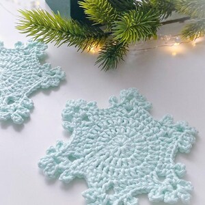May include: Two light blue, crocheted snowflake coasters. The coasters are on a white surface, with a green gift box and a sprig of evergreen in the background. The coasters have a detailed, openwork design.