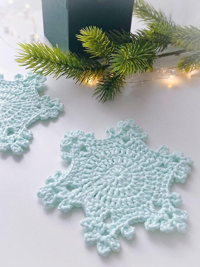 May include: Two light blue, crocheted snowflake coasters. The coasters are on a white surface, with a green gift box and a sprig of evergreen in the background. The coasters have a detailed, openwork design.