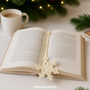 May include: An open book with a cream-colored crocheted snowflake bookmark. The bookmark is made of a cream-colored yarn and is placed between the pages. A cup of coffee and a Christmas tree are in the background.