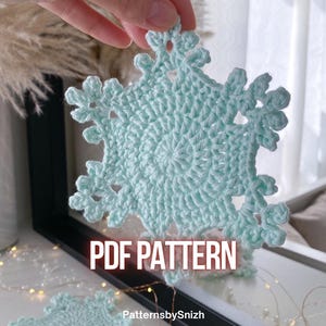 May include: A crocheted snowflake in a light blue hue is held up in front of a mirror. The snowflake has a detailed, openwork design. The text "PDF PATTERN" is displayed in white. The Etsy shop name "PatternsbySnizh" is also visible.