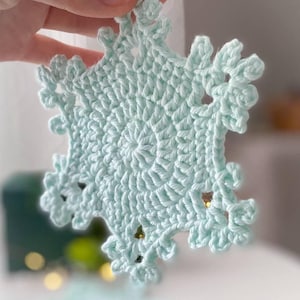 May include: A light blue, crocheted snowflake ornament. The snowflake has a detailed, openwork design with delicate, pointed edges. The ornament is held up, showcasing its intricate texture and shape. The snowflake is approximately 4 inches in diameter.