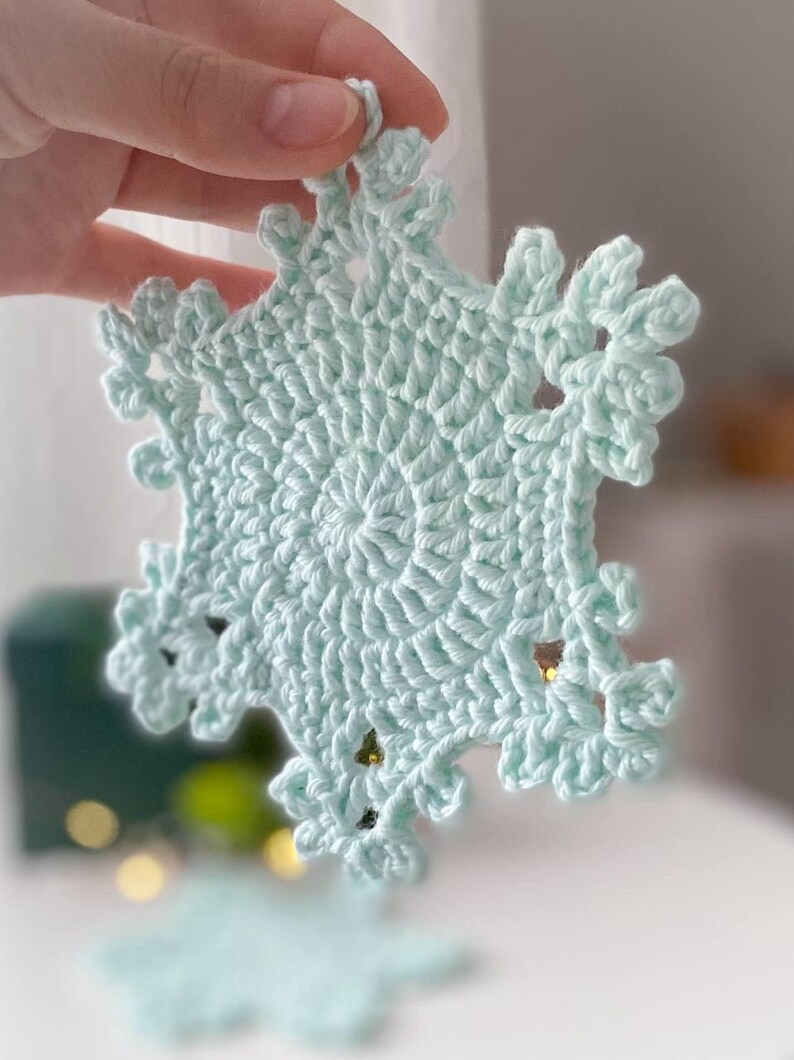May include: A light blue, crocheted snowflake ornament. The snowflake has a detailed, openwork design with delicate, pointed edges. The ornament is held up, showcasing its intricate texture and shape. The snowflake is approximately 4 inches in diameter.