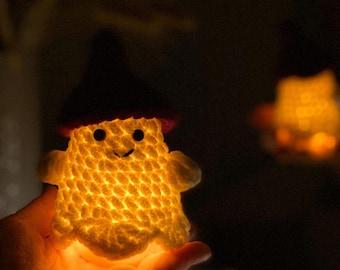 Puede incluir: Un fantasma brillante de ganchillo con una cara sonriente y un sombrero de bruja. La decoración de Halloween hecha a mano está iluminada desde dentro, creando un brillo cálido. El texto "PatternsbySnizh" es visible en la parte inferior.