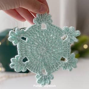 Könnte beinhalten: Eine gehäkelte Schneeflocke in einem sanften, pastellgrünen Farbton. Die Schneeflocke hat ein sechszackiges Design mit durchbrochenen Details und dekorativen Kanten. Der Text "PatternsbySnizh" ist unten rechts sichtbar.