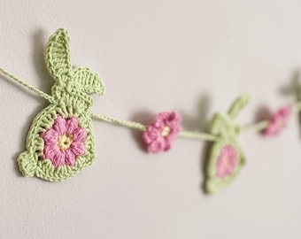 May include: A crocheted garland featuring green bunny shapes with pink flower centers and pink flowers. The garland is strung on a light green yarn and is displayed against a neutral wall. The text "PatternsbySnizh" is visible.