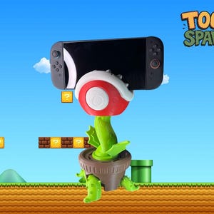 Op de afbeelding: Een nieuwe telefoon- of tablethouder in de vorm van een Piranha Plant uit de Super Mario Bros. videogame-serie. De houder is rood, wit en groen, met een apparaat met zwart scherm in zijn kaken. De achtergrond toont een cartoonlandschap.