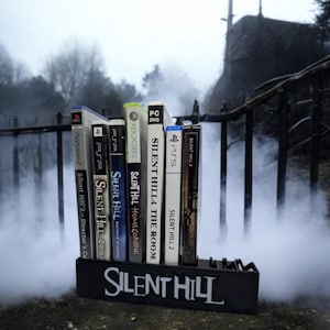 Może przedstawiać: Kolekcja pudełek z grami wideo Silent Hill, w tym wersje na PlayStation 2, PSP, Xbox 360 i PC DVD, umieszczona w czarnym uchwycie z napisem "Silent Hill". Scena rozgrywa się na zewnątrz, z mgłą i mrocznym, złowieszczym tłem.