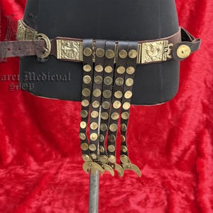 Puede incluir: Un cinturón de cuero marrón con detalles metálicos dorados, que incluyen placas decorativas y remaches. El cinturón presenta tiras de cuero colgantes con tachuelas doradas y piezas metálicas decorativas en la parte inferior. El cinturón se exhibe en un maniquí negro.