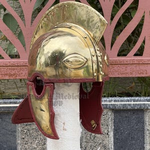 K&ouml;nnte beinhalten: Ein polierter r&ouml;mischer Helm aus Messing mit einem Nackenschutz aus rotem Leder. Der Helm hat einen markanten Stirnschutz und Wangenteile mit dekorativen Nieten und einer strukturierten Oberfl&auml;che. Der Helm wird auf einem wei&szlig;en zylindrischen St&auml;nder ausgestellt.