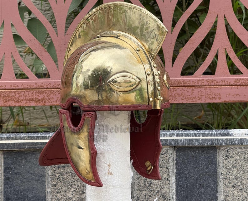 K&ouml;nnte beinhalten: Ein polierter r&ouml;mischer Helm aus Messing mit einem Nackenschutz aus rotem Leder. Der Helm hat einen markanten Stirnschutz und Wangenteile mit dekorativen Nieten und einer strukturierten Oberfl&auml;che. Der Helm wird auf einem wei&szlig;en zylindrischen St&auml;nder ausgestellt.