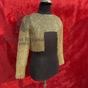 Könnte beinhalten: Ein goldfarbenes Kettenhemd mit langen Ärmeln, präsentiert auf einer schwarzen Schaufensterpuppe. Das Hemd besteht aus kleinen, ineinandergreifenden Metallringen. Der Hintergrund ist ein strukturiertes rotes Gewebe.