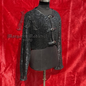 May include: A black chainmail jacket with long sleeves and leather accents. The jacket is displayed on a mannequin against a red backdrop. The jacket has three leather straps with silver buckles. The product is from Margaret Medieval Shop.
