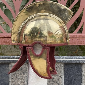 May include: A brass Roman helmet with a polished finish. The helmet features a curved top, eye guards, and cheek pieces with a red leather trim. The helmet is a historical replica.