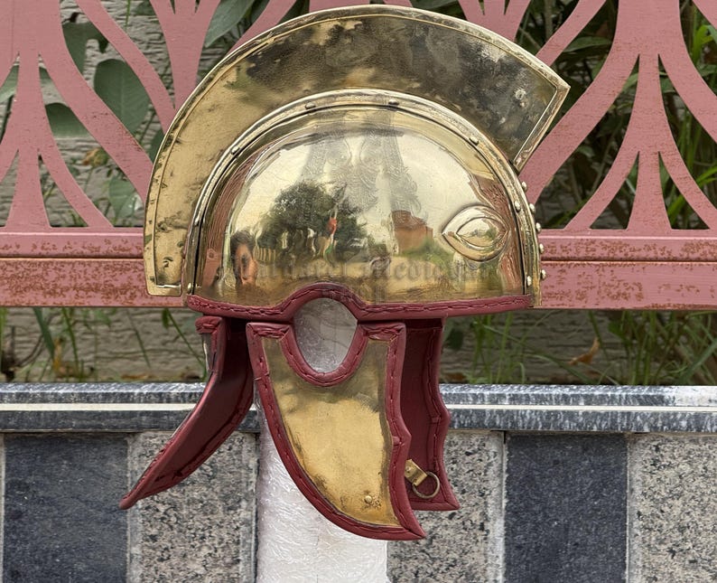 K&ouml;nnte beinhalten: Ein r&ouml;mischer Helm aus Messing mit poliertem Finish. Der Helm hat einen gebogenen Oberteil, Augenschutz und Wangenplatten mit roter Lederausstattung. Der Helm ist eine historische Nachbildung.