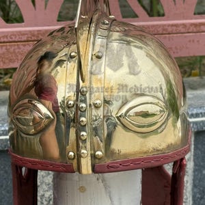 K&ouml;nnte beinhalten: Ein polierter Messinghelm mit reflektierender Oberfl&auml;che. Der Helm hat augenf&ouml;rmige Aussparungen und ist mit rotem Leder verkleidet. Die Oberseite hat einen vertikalen Grat und ist mit Nieten verziert. Historische Replik.
