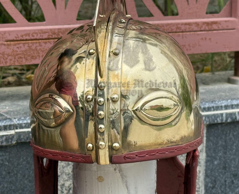 K&ouml;nnte beinhalten: Ein polierter Messinghelm mit reflektierender Oberfl&auml;che. Der Helm hat augenf&ouml;rmige Aussparungen und ist mit rotem Leder verkleidet. Die Oberseite hat einen vertikalen Grat und ist mit Nieten verziert. Historische Replik.