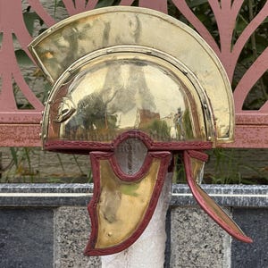 K&ouml;nnte beinhalten: Ein polierter r&ouml;mischer Helm aus Messing mit gew&ouml;lbter Oberseite und roten Lederakzenten. Der Helm hat Wangenklappen und einen dekorativen Kamm, der historische Handwerkskunst zeigt. Der Helm ist eine Nachbildung antiker Milit&auml;rhelme.